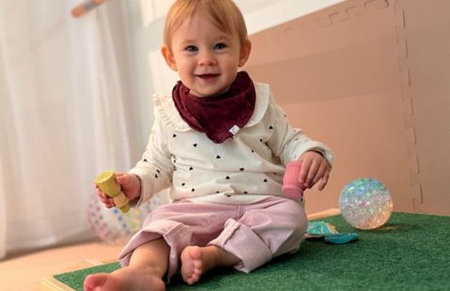 Baby sitzt auf einem Krabbelpodest und hält in der Hand Holzspielzeug